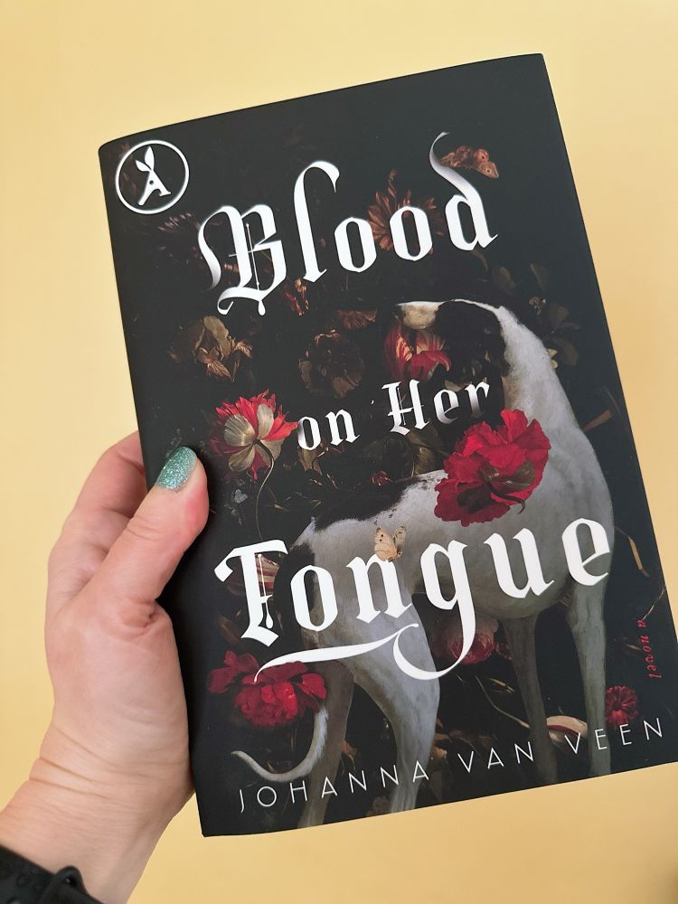A hand holding the hardcover copy of Blood on Her Tongue by Johanna Van Veen