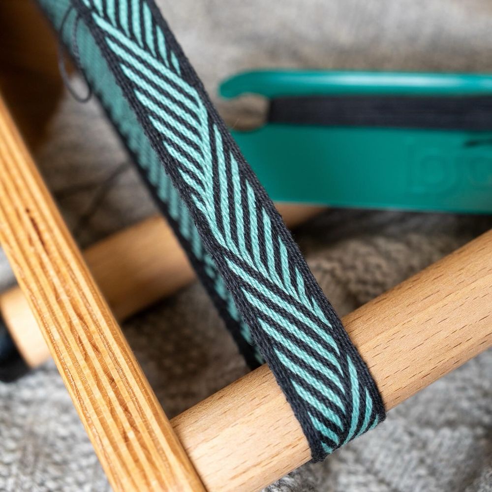 A 2cm wide band on an inkle loom. The design features diagonal stripes of black and aqua which traverse the band in triangular blocks. It’s not quite a symmetrical design. Each alternate triangle on the right is bordered either with the main or background colour. 
