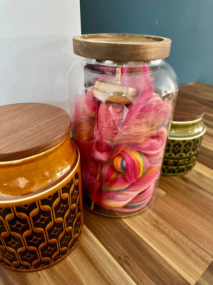 A glass jar full of colourful fibre and a spindle on a kitchen counter 