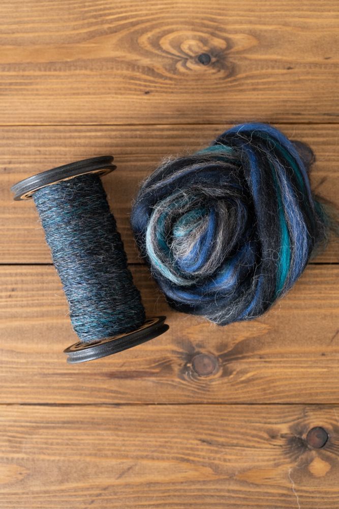 Mineralogy ‘Blue Slate’, a top containing royal blue, teal, black and white which spins up to a complex dark blue. Pictured in tops form and handspun singles on a bobbin. 