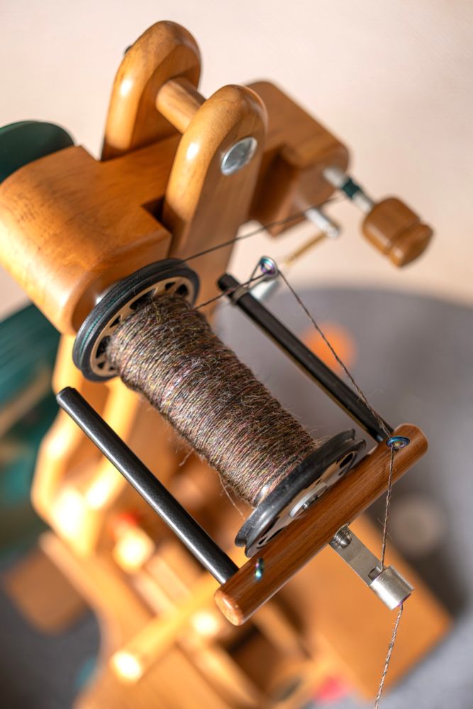 A Majacraft Suzie Pro spinning wheel fitted with the lace flyer. On it is a half-full bobbin of a complex, green-leaning brown yarn  