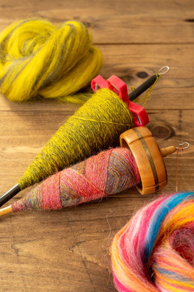 Two drop spindles. One 3D printed with a pink whorl and bright green yarn wound in a standard cop; one wooden with a brass inlay around the rim and wound with a mysterious mixture of bright pink and… all the colours! Displayed in a fancy-looking cop where each make is visible in the criss-crossed pattern. The fibre they’re spun from sits beside them.