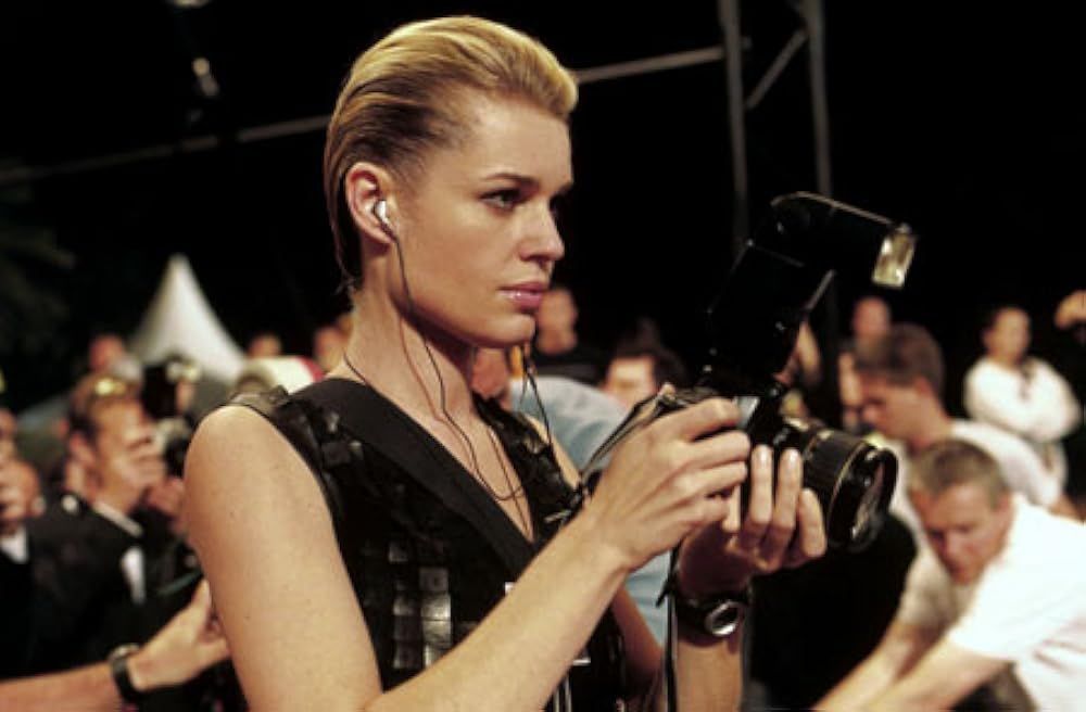 Rebecca Romijn in FEMME FATALE in a crowded room with slicked back hair, a black leather top, holding an SLR camera