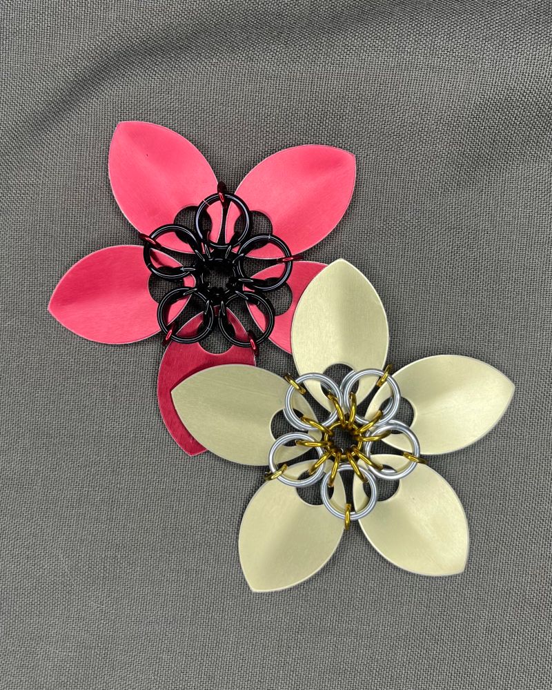 Two Scale Mail flowers, intended for use as holiday ornaments and pendants. This is my second size, the larger of the single layer flowers. Each is made from five large scales connected by 16 and 18 gauge rings. The first is made from red scales connected by black rings, the second is made from gold scales connected by gold and silver rings, meant to evoke a holiday star. They are sitting together on a neutral fabric background.