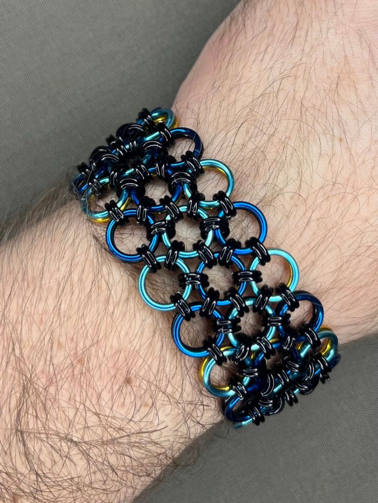 One side of a reversible chainmail cuff bracelet in the Japanese 12-in-2 weave. The three row bracelet is made from anodized aluminum rings, with alternating diagonal lines of royal blue and sky blue, all connected by smaller black rings. Hints of the black and yellow other side can be seen. Modelled by myself against a neutral fabric background.