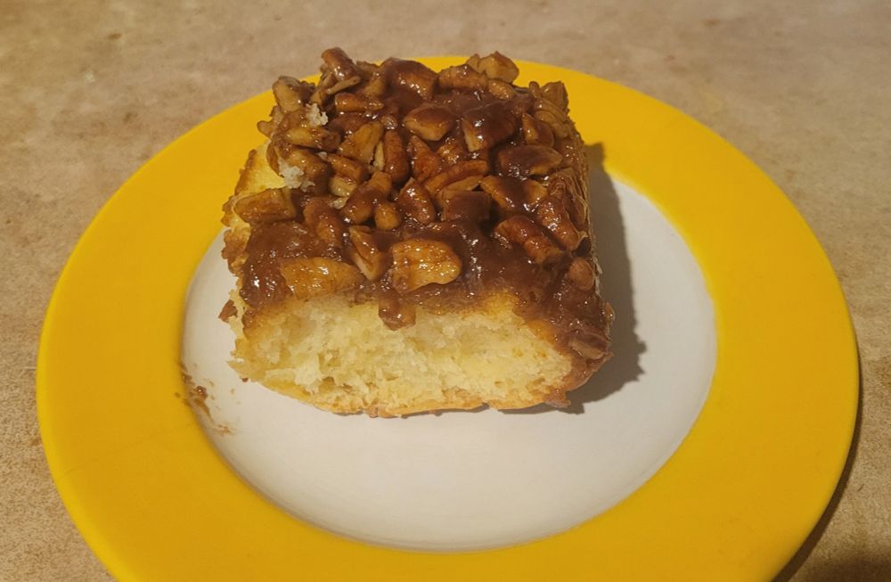 One sticky bun on yellow and white plate. Fluffy bun with a heap of pecans on it with a caramel-like maple sauce. 