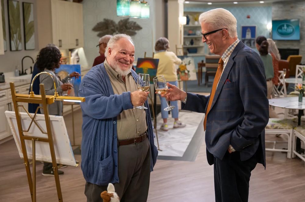 Two older men of different races, one tall wearing a suit and glasses and one wearing a thin cardigan over collared shirt and pants, toast with wine glasses in a large open kitchen where people are gathered at various paintings making art.