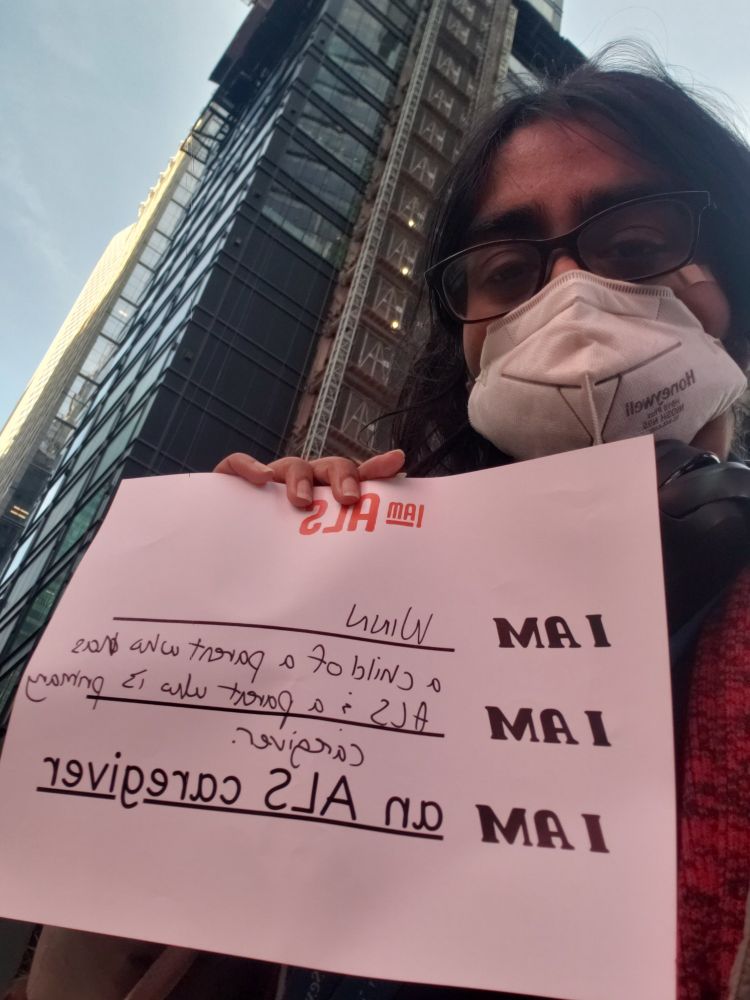 A masked person of color holds an i AM als caregiver sign introducing themselves and the person they're caregiving for