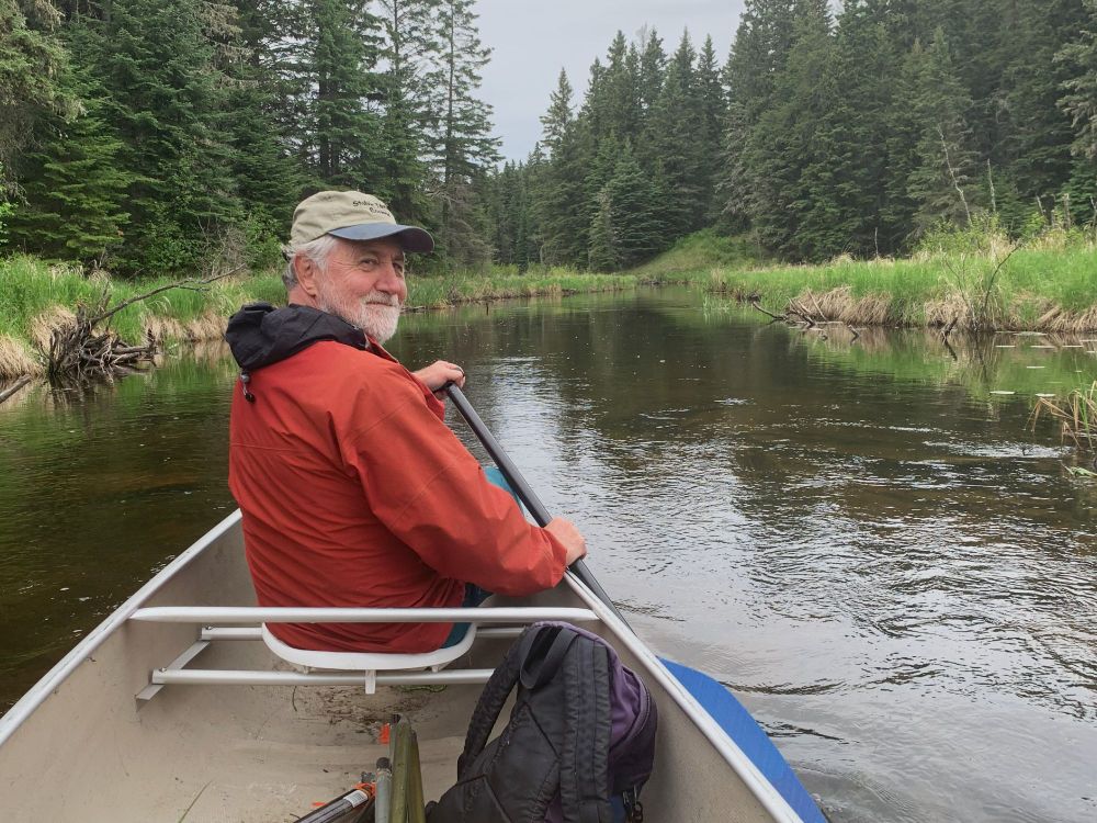 Dr. Keith A. Hobson, an older man with a white beard, paddles a canoe along a scenic river