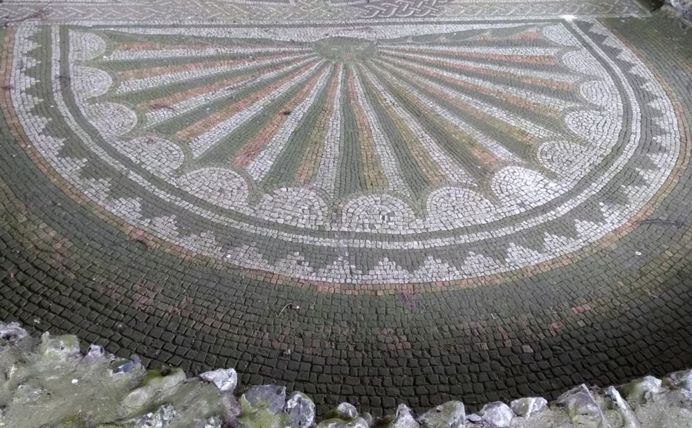 Part of a Roman mosaic showing an apse with a radial design in the form of a shell, with the segments coloured in dark grey, red and white/cream. The shell's hinge is at the top of photograph and takes the form of a small cat-like mask. There is a border of stepped triangles and a wider plain outer border. In the immediate foreground are the remains of the wall around the apse.