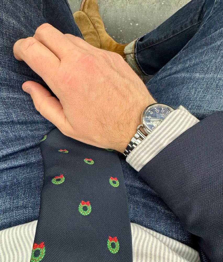 Looking down at a striped oxford shirt and navy blue tie with green wreaths on it, along with a hand and wrist wearing a watch 