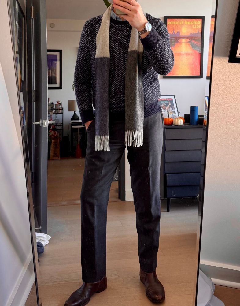 Man standing in front of the mirror, wearing dark gray wool pants, dark brown leather boots, a navy blue sweater with a white sorta birdseye pattern, and an off-white scarf with dark gray on one end and dark blue on the other 
