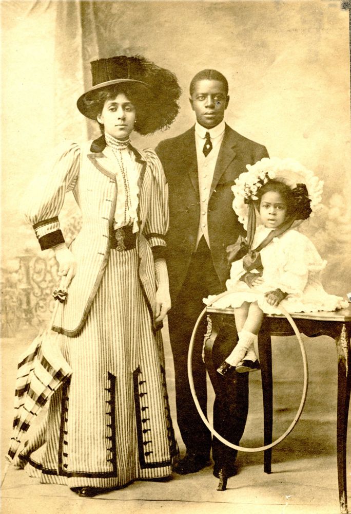 A formal portrait of the Taylor family. On his left, Taylor's wife Daisy Victoria Morris poses with a hand at her hip and dons a large feathered top hat. At his right, Taylor's daughter Syndey is dressed in a frilly gown and sits atop a table. A hoop is draped over her knees. Taylor stands proudly in the center, dressed in a black suit.