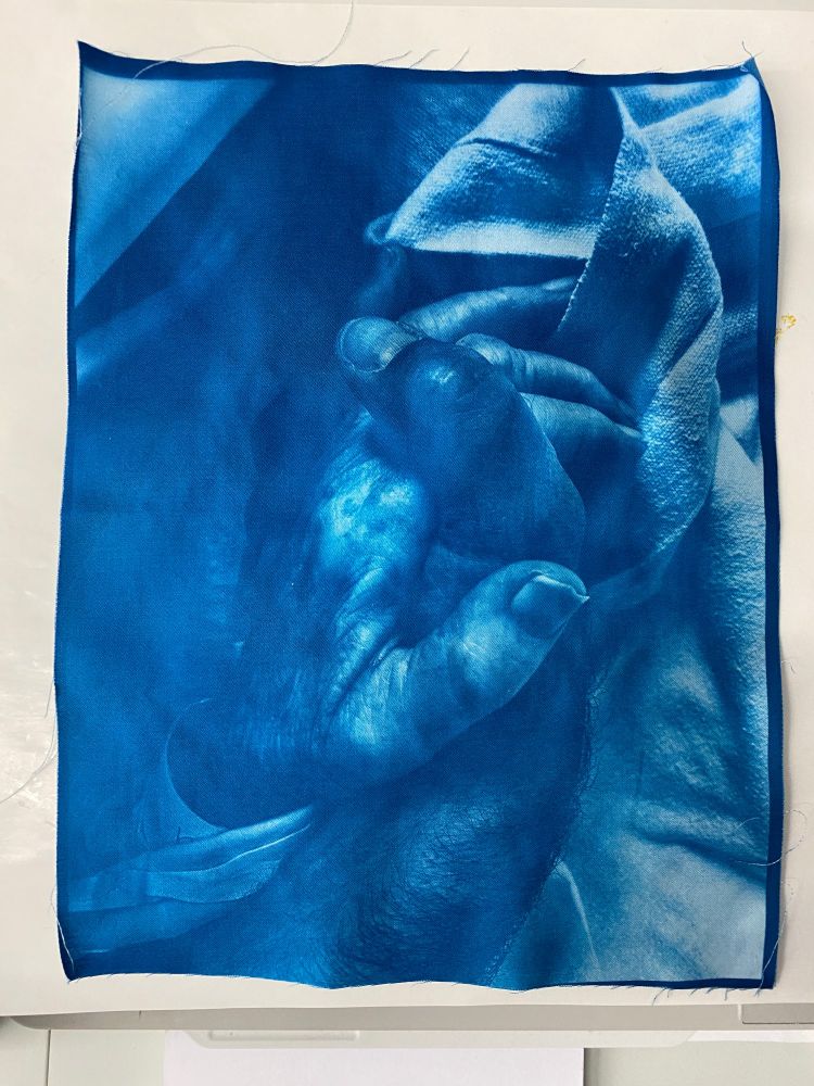Colour photo of a fabric cyanotype print depicting my hand holding the hand of my mother, who was in the hospital after breaking her hip. Living with dementia, she was unable to walk again. 