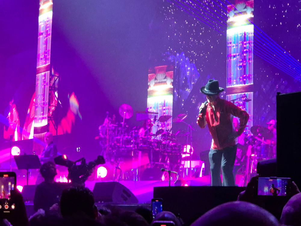 Jay Kay van Jamiroquai op het podium in de Ziggo Dome in een van zijn outfits van de avond, cowboyhoed en rood sportjasje. Op de achtergrond prachtig kleurrijke visuals.