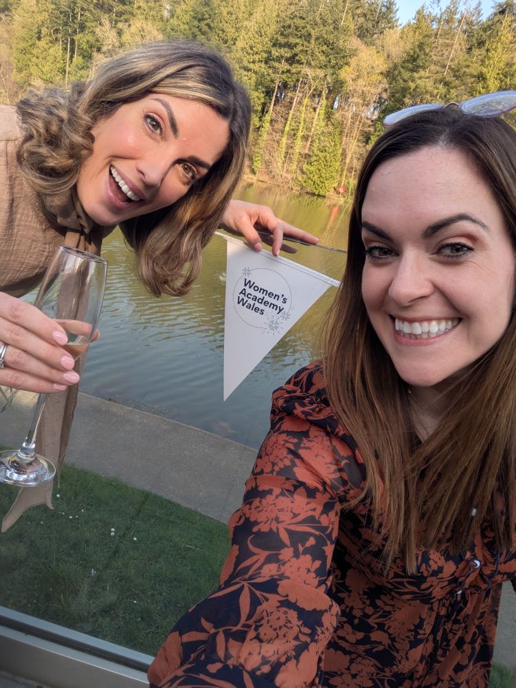 Andrea and Emma with champagne in front of bunting and a lake 