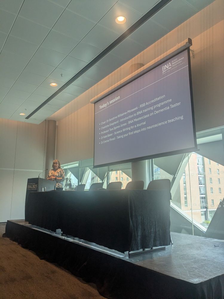 Sandrine Williams a white woman stood behind a lecturn and in front of a PowerPoint slide in a conference room