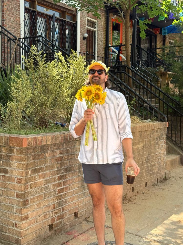 Man wearing a striped button up shirt, blue shorts, and a yellow hat holds sunflowers and an iced coffee, this time smiling with the flowers close to his face. 