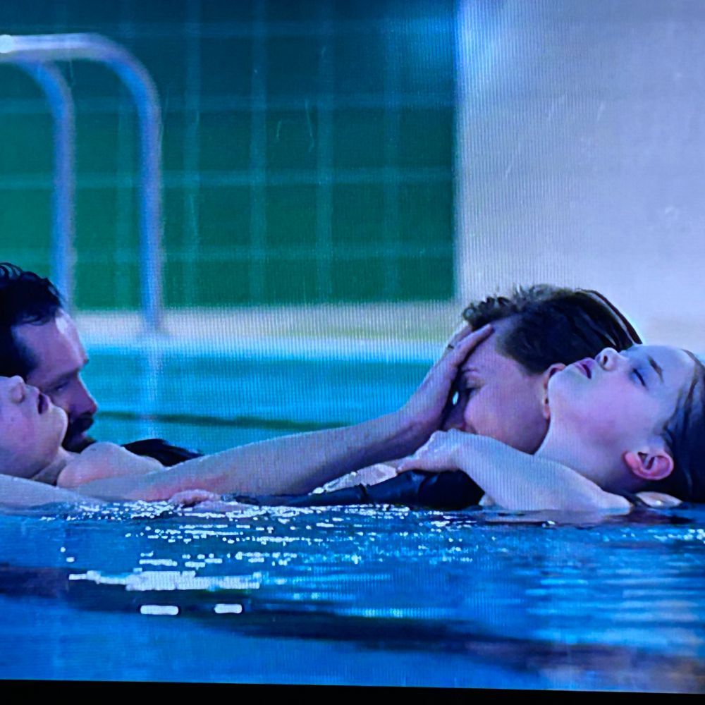 une famille dans une piscine, le père et la mère portent chacun un enfant qui dors dans l'eau. Le père caresse le front de la mère en pleur 