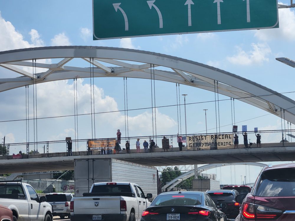A sea of cars in traffic below the Dunlavy St bridge over I-69N with protesters holding signs like RESIST RACIAL GERRYMANDERING.