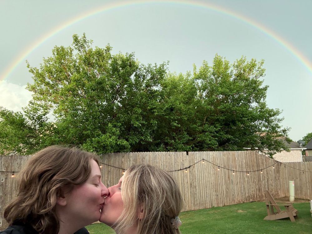 A photo of two women kissing under a rainbow during pride month 