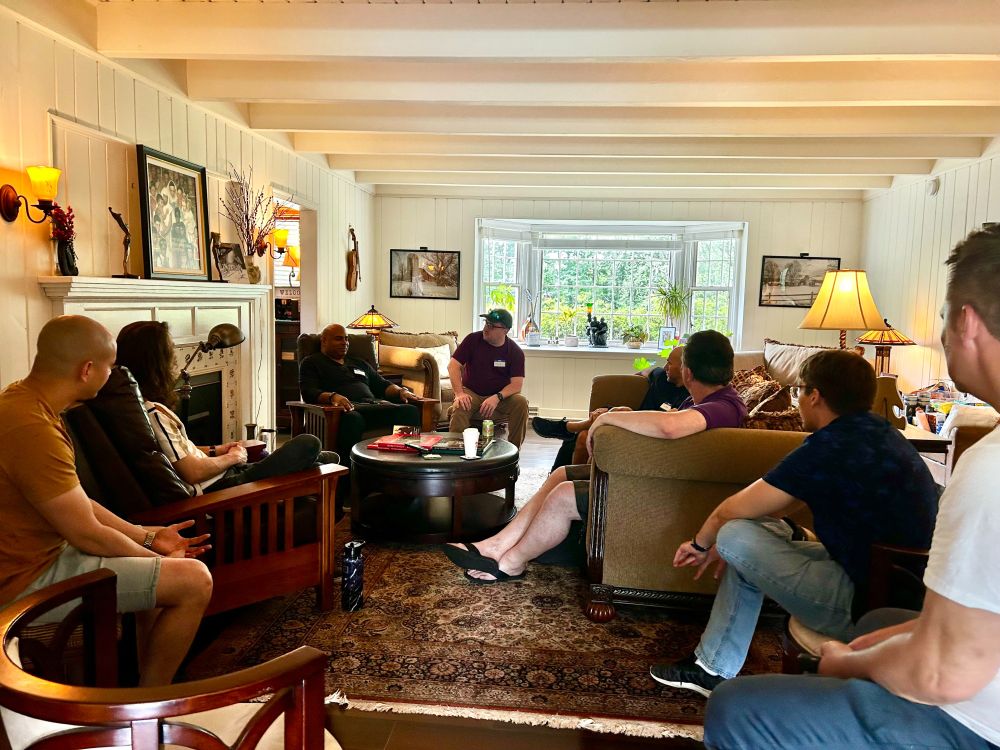 NJTC group members around a coffee table in The Ampersand Inn living room, all looking intently at Garth, who is speaking