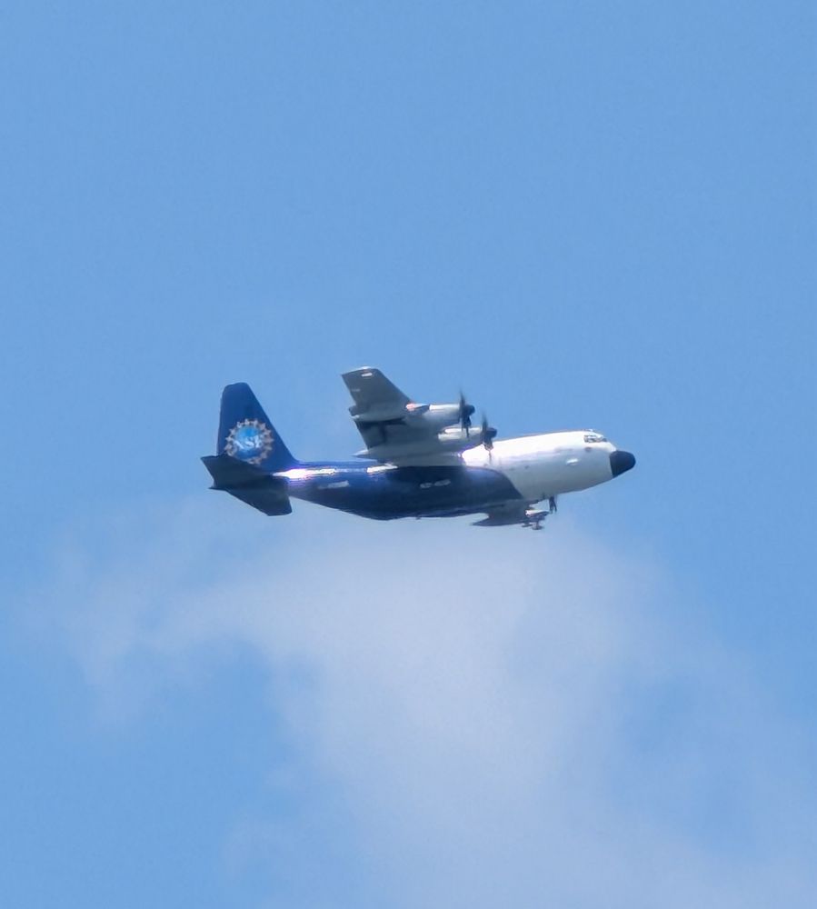 A C-130 aircraft wearing the emblem of the NSF flying among a blue sky.