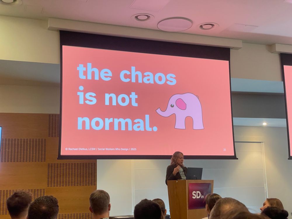 Photo of Rachael speaking. The slide behind her says "the chaos is not normal." With an illustration of a pink elephant.