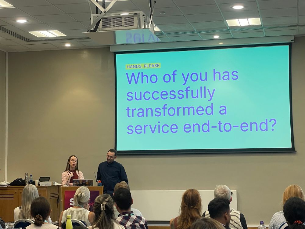 Photo of Kara presenting a workshop with Martin Jordan. The slide behind them says "HANDS, PLEASE
Who of you has successfully transformed a service end-to-end?"