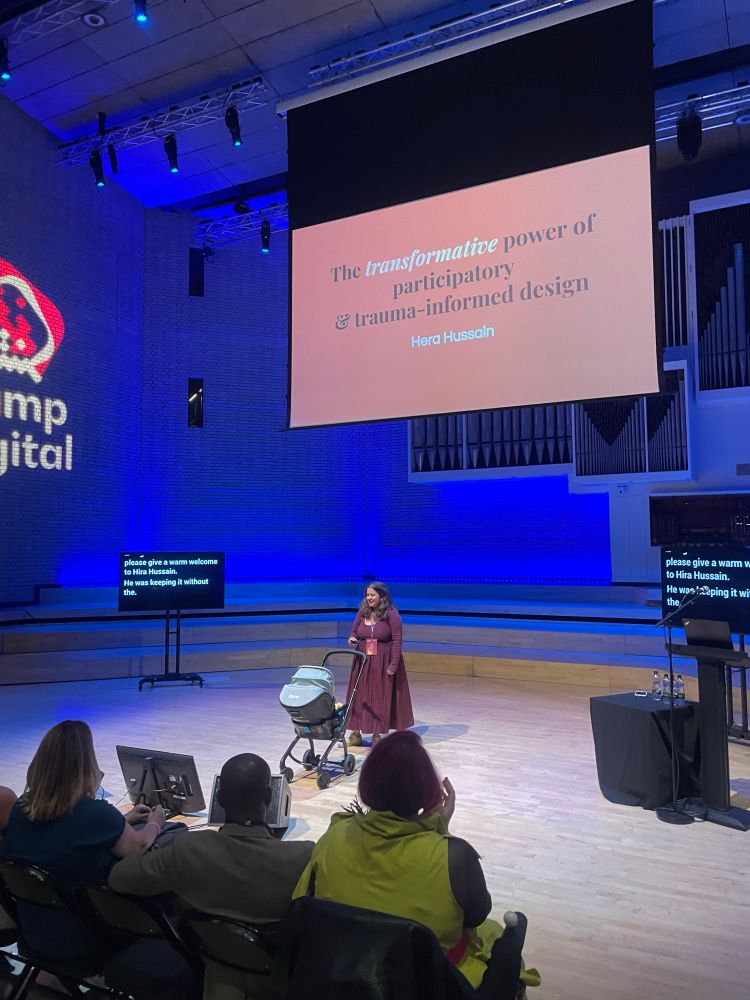 Hera on stage with a pram, the slide says "The transformative power of participatory and trauma-informed design" and the wall says "Camp digital"