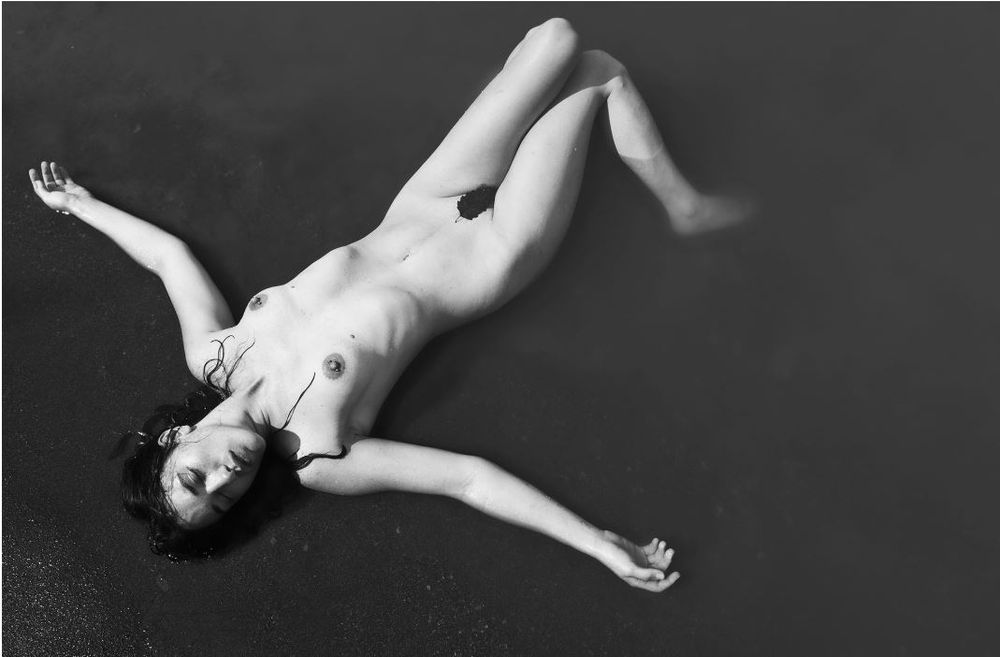 Washed Ashore photo taken by Mynameisaldus of traveling model Sirena Wren. She is laying on a beach with her arms out like she was washed ashore on the beach.. The lighting shows great depth in shadows from her skin to the shades of grays. 