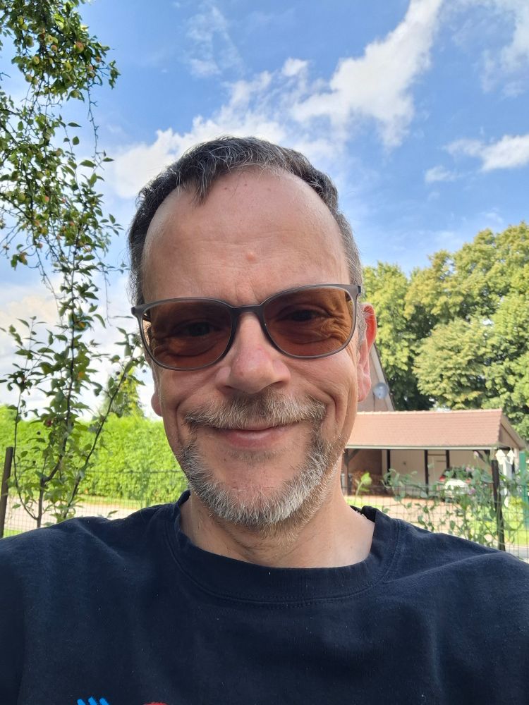 Das Bild zeigt einen Mann mit grauem Bart und Sonnenbrille, der nach oben schaut. Er trägt ein dunkelblaues Shirt mit einem kleinen Logo. Im Hintergrund ist ein klarer blauer Himmel mit einigen Wolken zu sehen, sowie grüne Bäume und Sträucher. Es scheint ein sonniger Tag an einem Ort mit Natur zu sein.