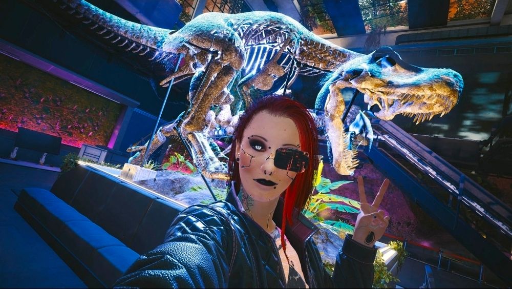 Photo of V from Cyberpunk 2077. The red-haired character is giving the peace sign, and posing in front of a hologram of a T. Rex skeleton