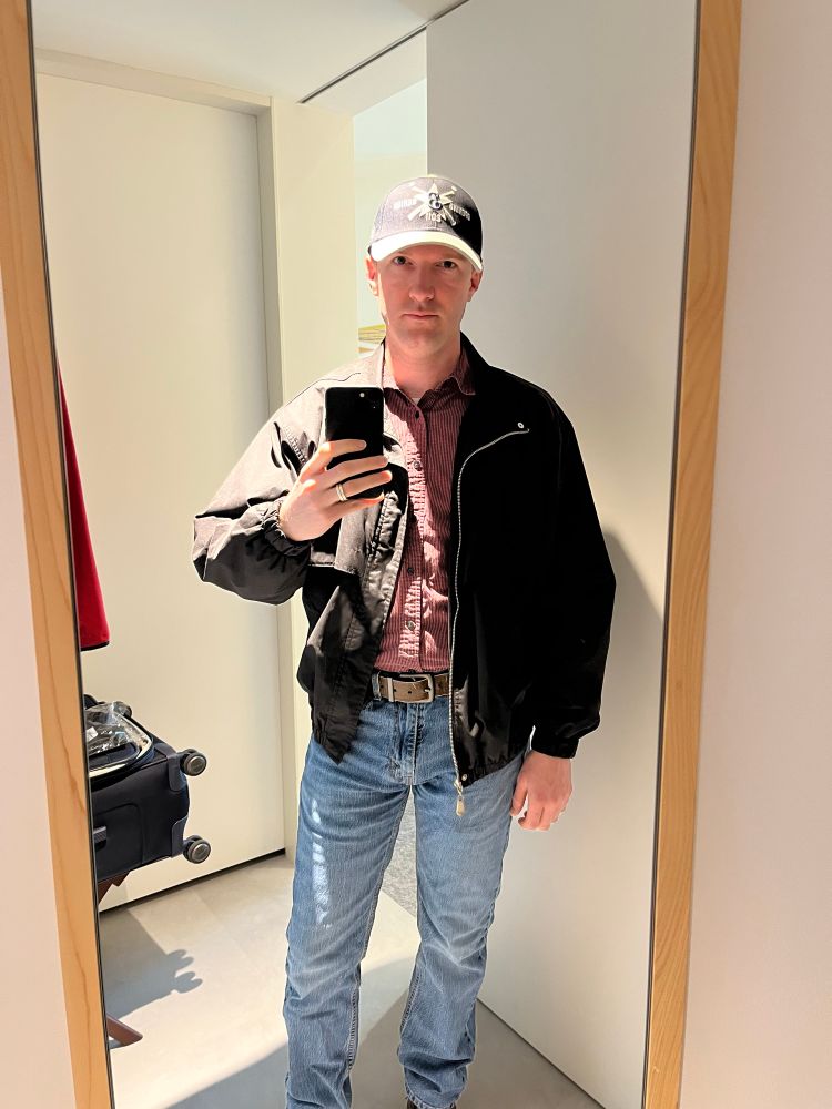 Guy in his late thirties wearing a button down shirt tucked into jeans with a belt, a windbreaker, hiking boots, and a 14 year old baseball cap.