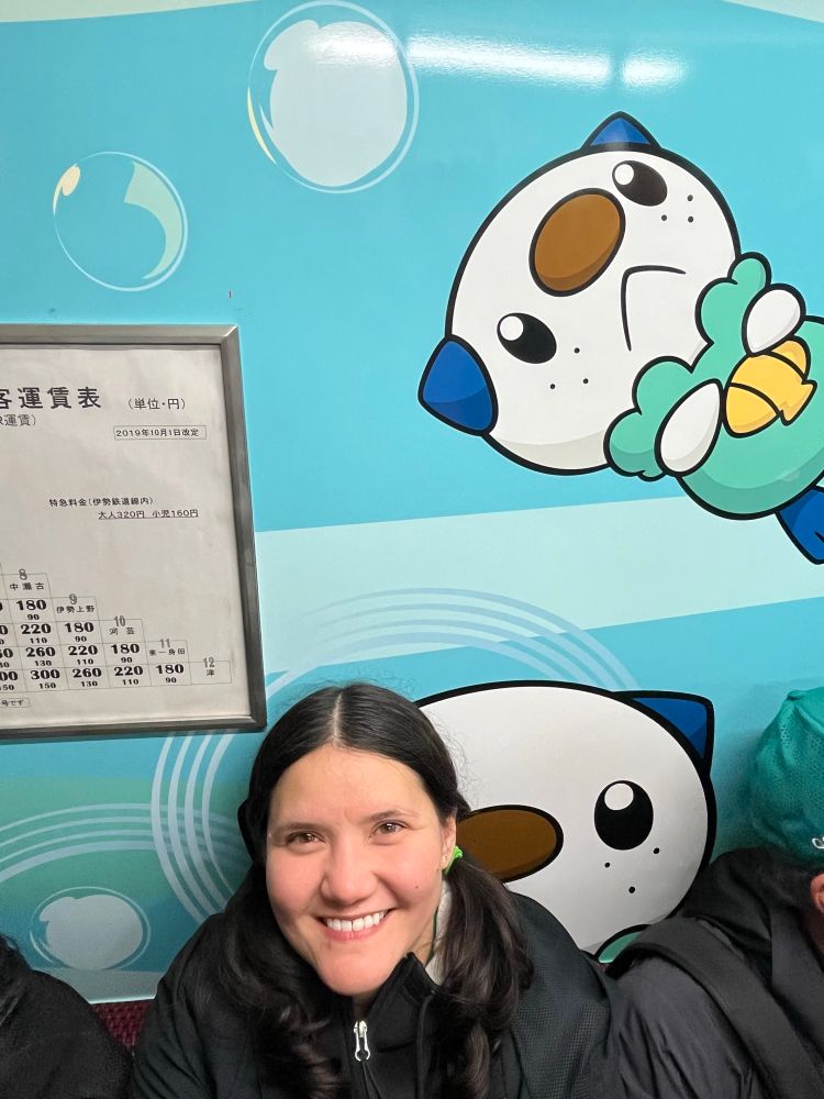 Molllie smiles, sitting inside a train. The inside walls of the train have a mural with the Pokémon, Oshawott. 