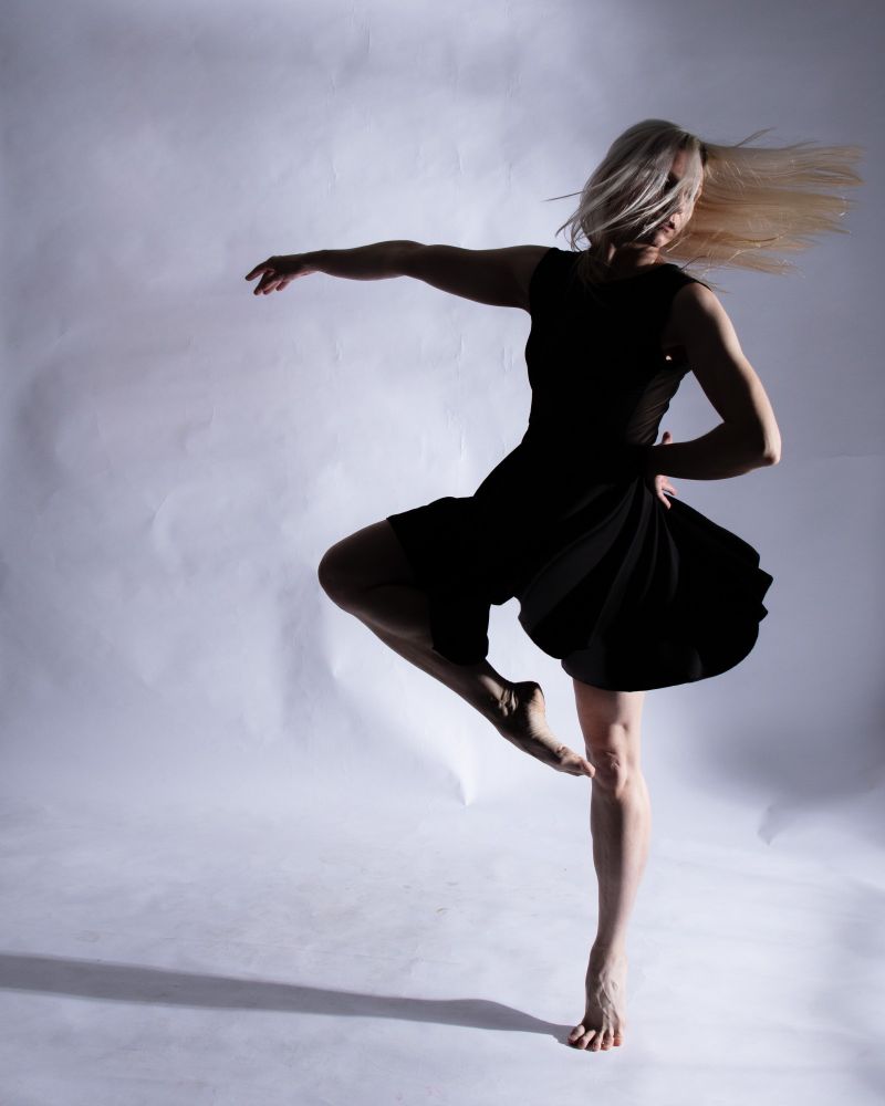 A dancer on her toes wearing a black dress against a white paper backdrop, in the middle of a pirouette, blonde hair catching the strong side light