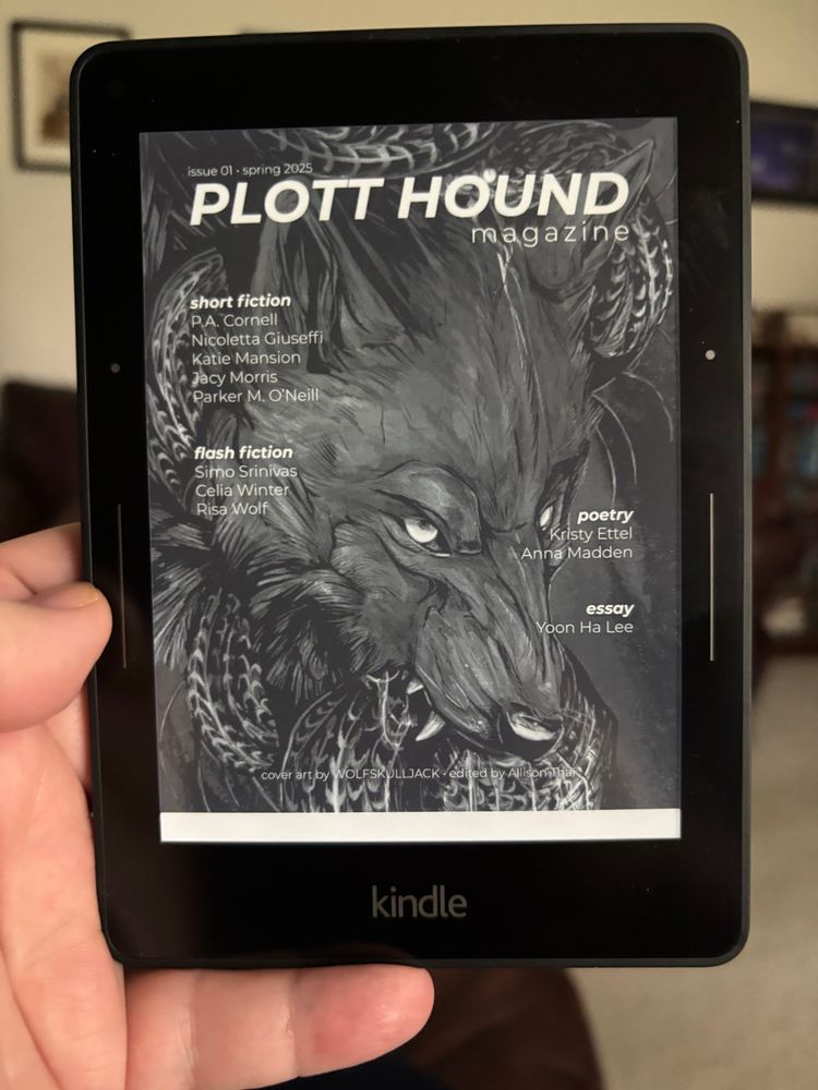 Picture of a Kindle Voyage ereader displaying the cover of Plotthound issue 1 which displays are of a toothy werewolf by @wolfskulljack, the contents of the magazine are typeset around the image. Edited by Allison Thai. 