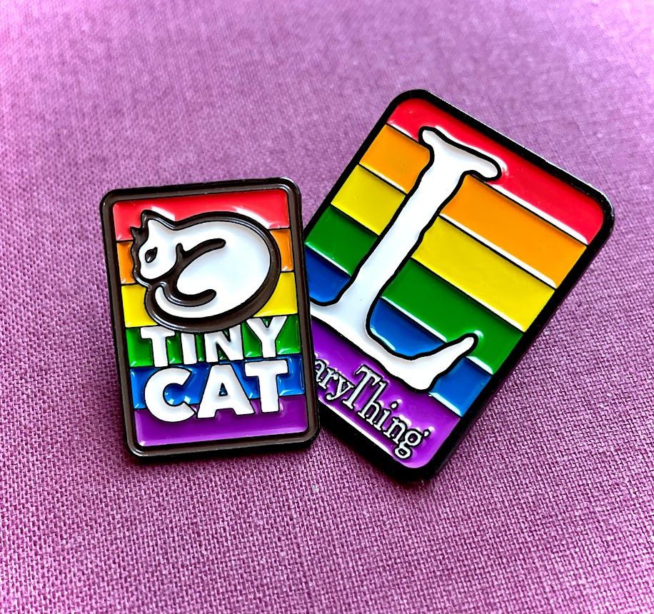 Photograph of two rainbow enamel pins, one showing the TinyCat logo and the word TinyCat, the other showing the LibraryThing logo and the word LibraryThing.