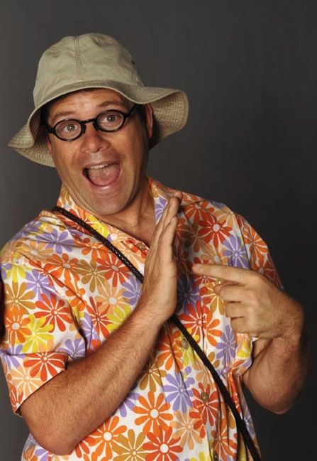 A color photograph of a light-skinned man wearing a loud Hawaiian shirt with random ten-petaled flower prints. He has a floppy hat, dark-rimmed glasses and a dark strap running from shoulder to hip. His mouth is open in an expression of excitement and he's pointing at his right palm with his left forefinger. The background is a neutral gray.