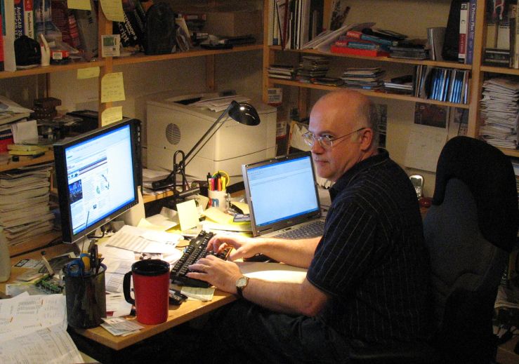 Color photograph of a balding middle-aged man sitting at a messy desk with several computer monitors. In the background are wooden shelves loaded with books, CDs and test equipment.