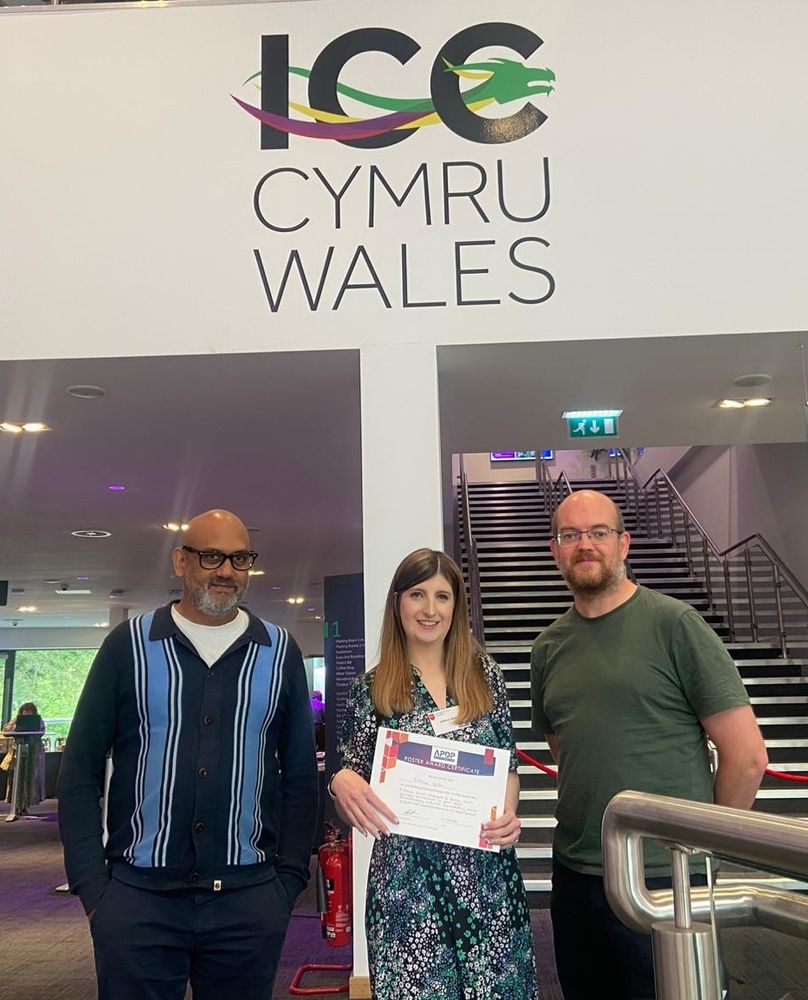 L-R Dr Mark Paul-Clark, Dr Jessica Bates, Dr Richard Hulse. Jess is holding her prize certificate for best science communication. They are standing in front of signage "ICC Cymru Wales".