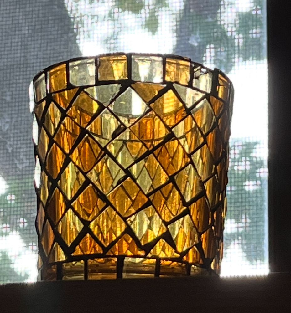 Small glass candleholder on a shelf in front of a screened window. Trees can be seen outside. It is daylight and light shows patterns on the various shades of amber diamond shaped pieces that are the main part of the glass, with rectangular at top and bottom. 