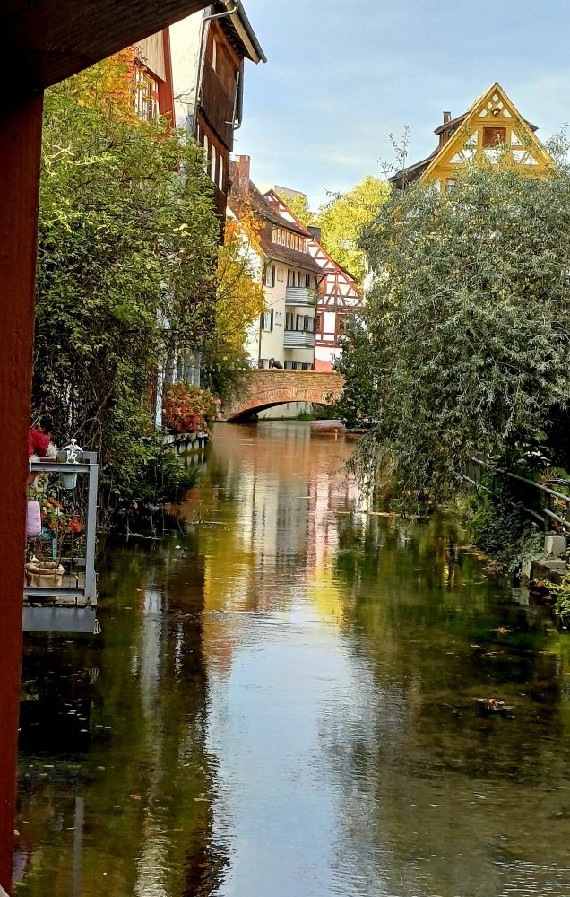 Ein ruhiger Fluss, eine alte Steinbrücke darüber, Häuser direkt am Fluss mit Balkonen, die über das Wasser ragen.