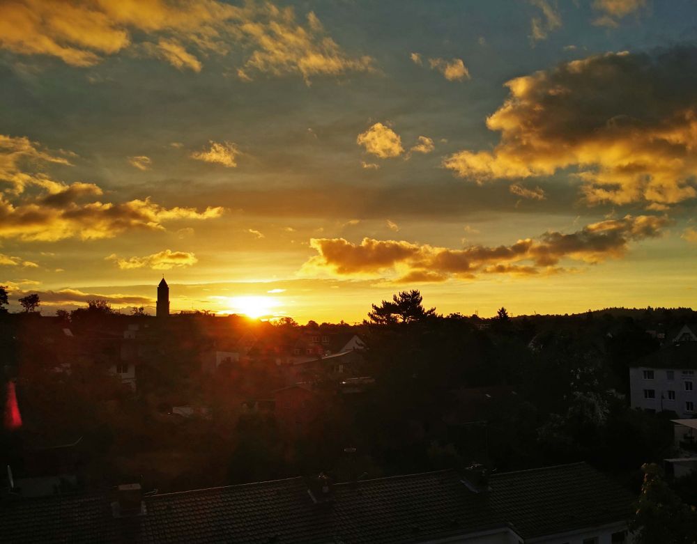 Sunrise over a city with a church and a tree in the background under a gold-coloured cloudy sky.