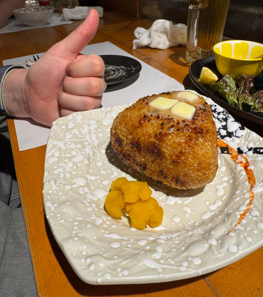 A grilled rice ball much larger than a man’s closed fist. Topped with butter. Thumbs up for scale. Japanese pickles on the side.