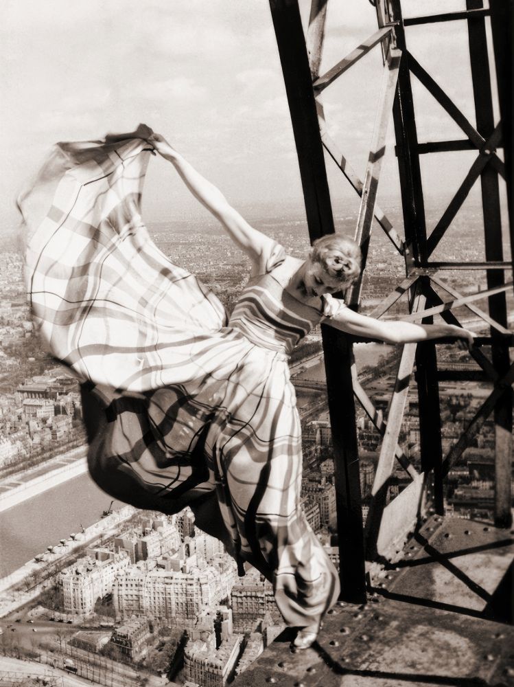 A black and white photograph, sepia tinted, of model Lisa Fonssagrives. She is on the Eiffel Tower, out on the framework of the tower, balancing on the edge of a drop several hundred feet to the city which is visible below her and stretching to the skyline. She has on a dress with a wide skirt and she is holding the hem of the skirt above her head, her arm outstretched so that a great expanse of fabric billows out over the city. It is a bright sunny day and she seems full of joy and courage.