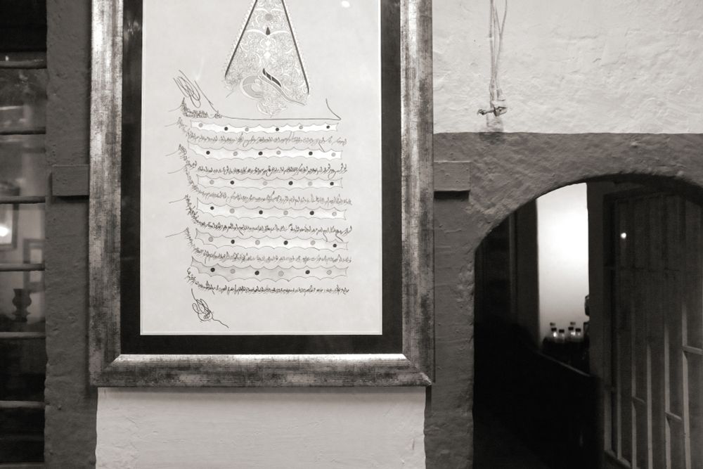 black and white photo of a section of wall, on the right there is a doorway into a darkened room. On the wall there is a piece of calligraphy, embellished with gold, in a gilt frame. The whole composition is an arrangement of rectangles.