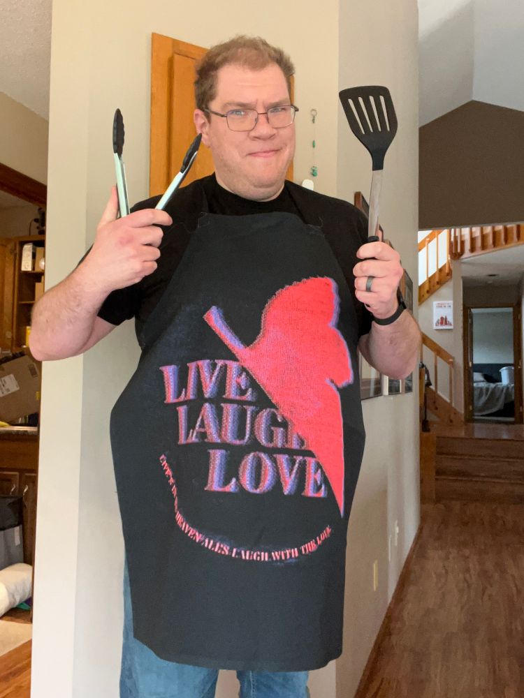 Shout wearing an apron with a parody of the NERV logo that reads, "Live, Laugh, Love: Live's in His heaven, all's Laugh with the Love"