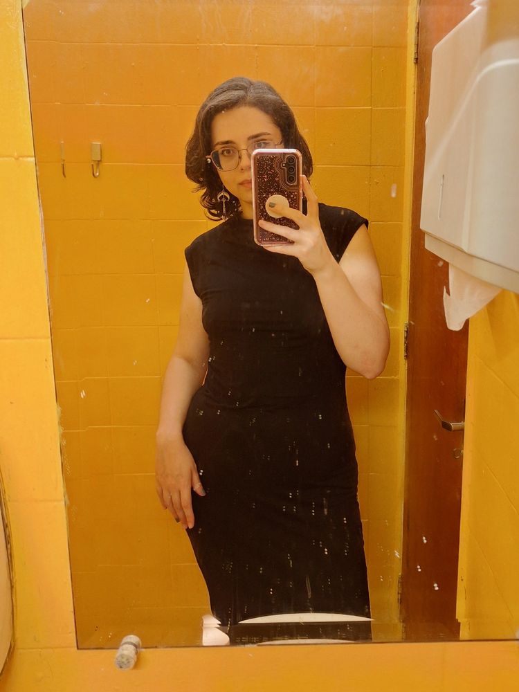 minha foto com um vestido preto na frente do espelho de um banheiro pequeno de restaurante, de paredes amarelas.