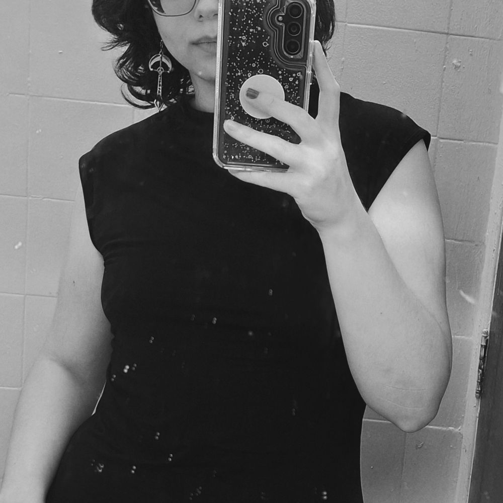 minha foto com um vestido preto na frente do espelho de um banheiro pequeno de restaurante, de paredes amarelas, mas em preto e branco